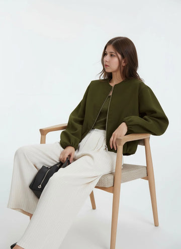 Woman sitting on a chair wearing an olive green jacket and white pants, holding a black bag.