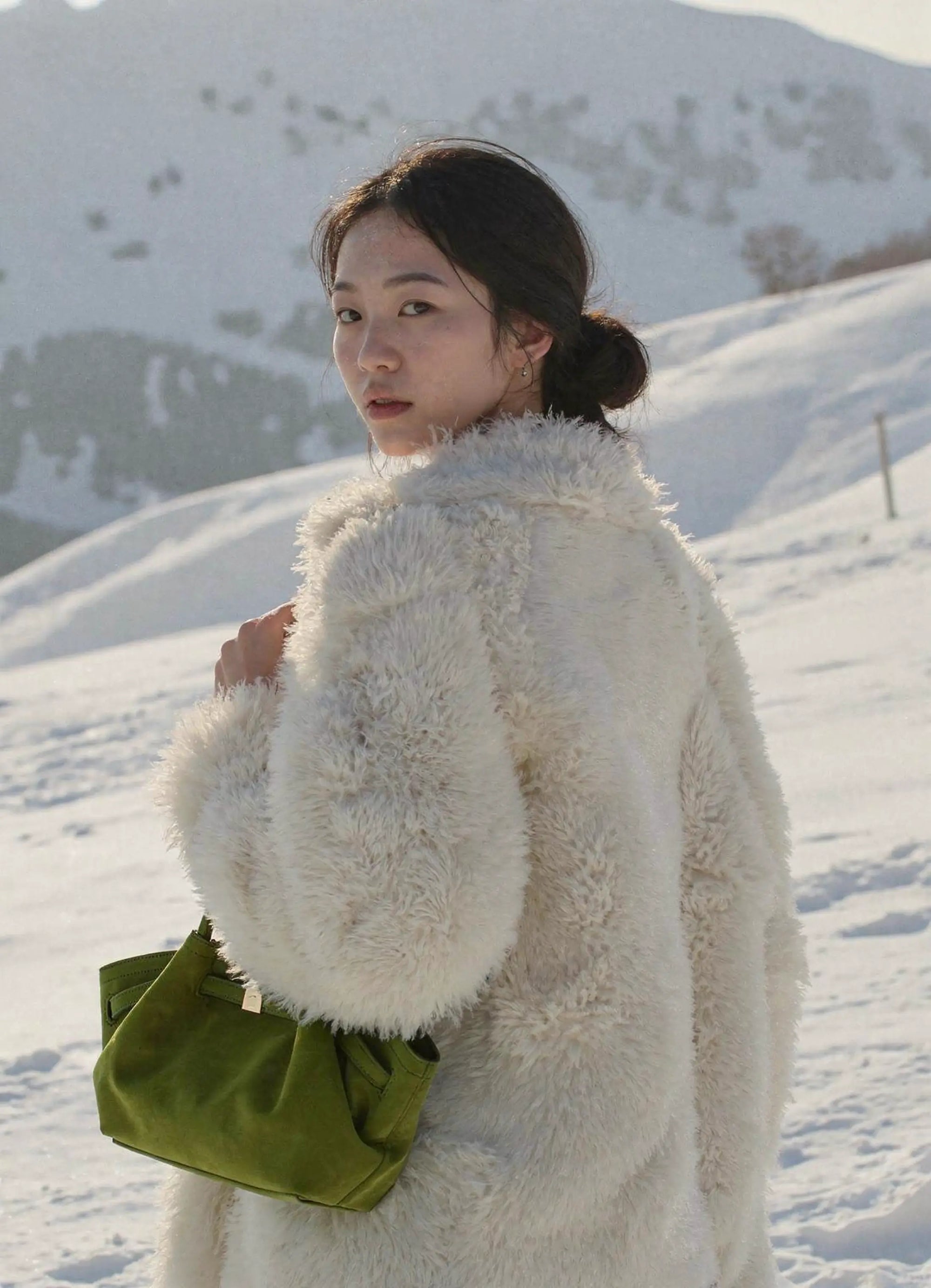 Woman in a white fluffy coat and green bag standing in a snowy landscape