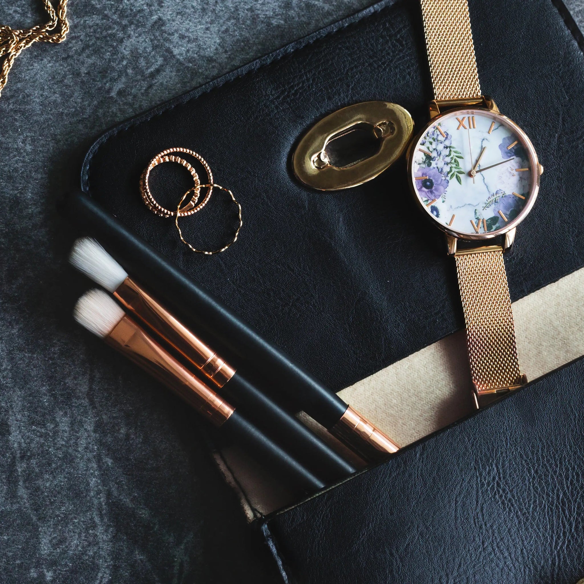 Watch, makeup brushes, and jewelry on a dark surface